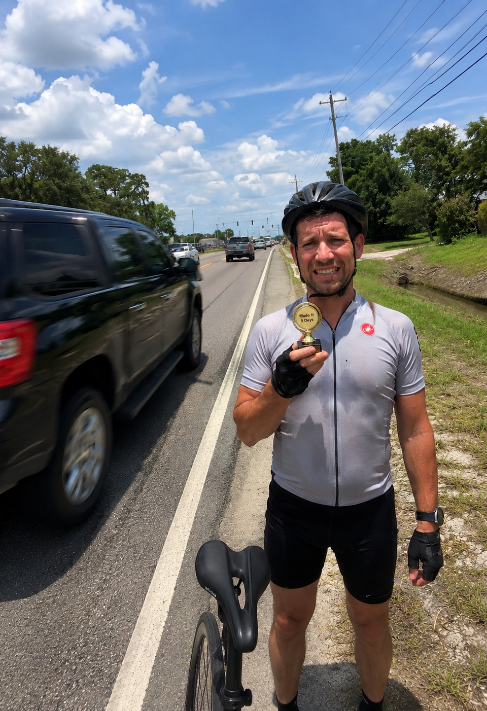 A sweaty cyclist standing beside a narrow bike lane on a busy road, holding a small “Made It 5 Days” trophy while cars and trucks pass close by.