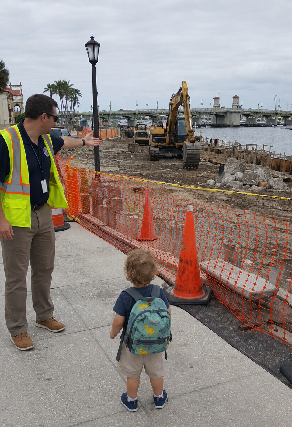 A toddler with a backpack stands beside orange construction fencing on the St. Augustine bayfront while a city worker gestures toward seawall construction near the Bridge of Lions.