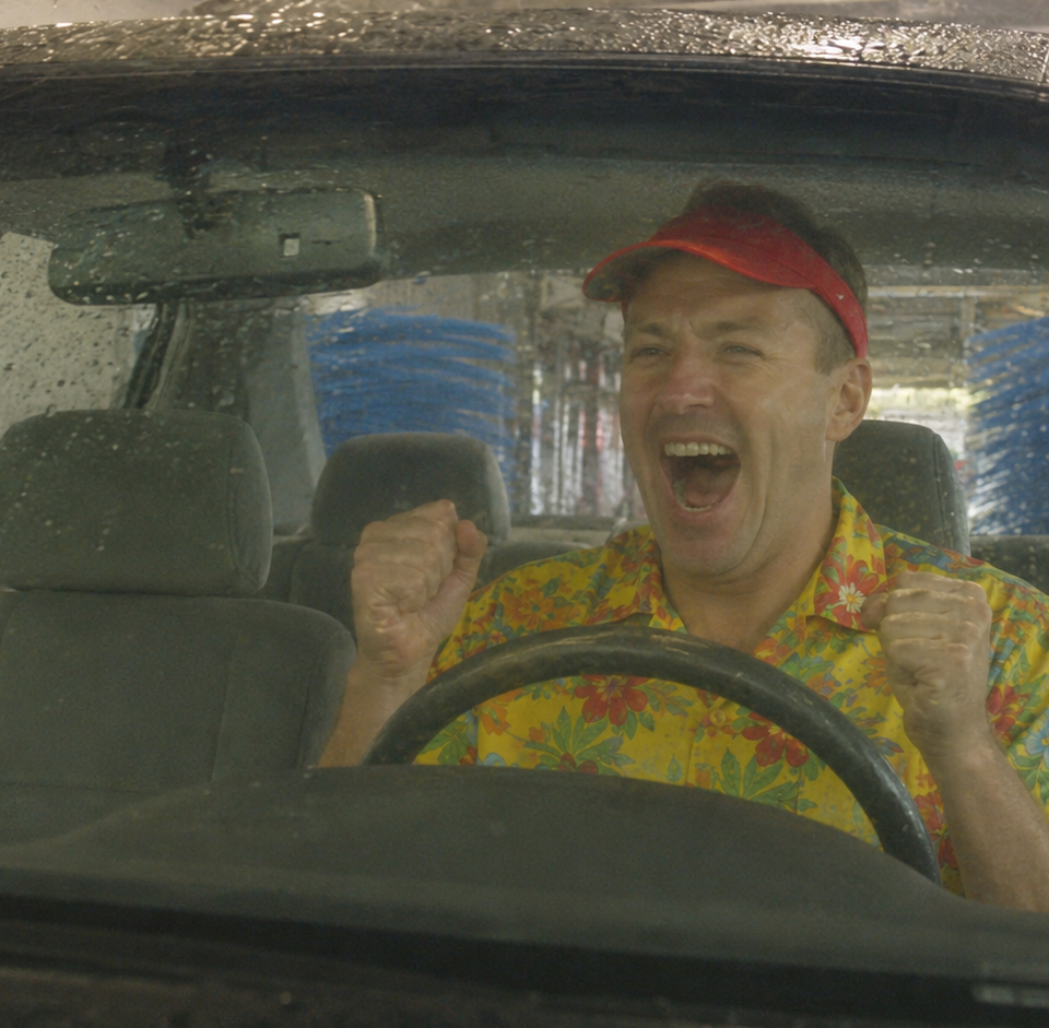 Excited man raises his fists inside a car as it moves through an automatic conveyor car wash.