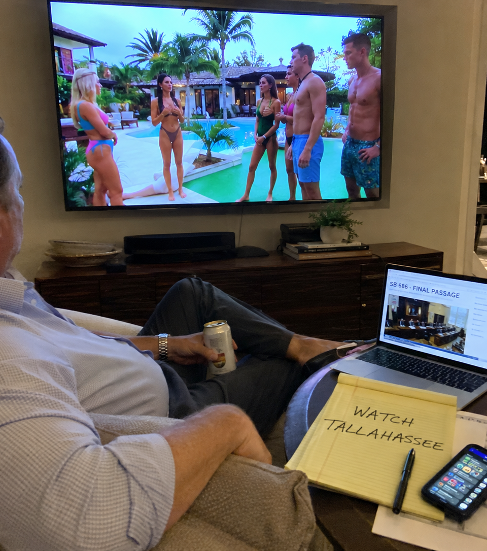 Satirical image of a fictional county lobbyist watching a tropical dating show on TV while ignoring a laptop displaying state legislative business.
