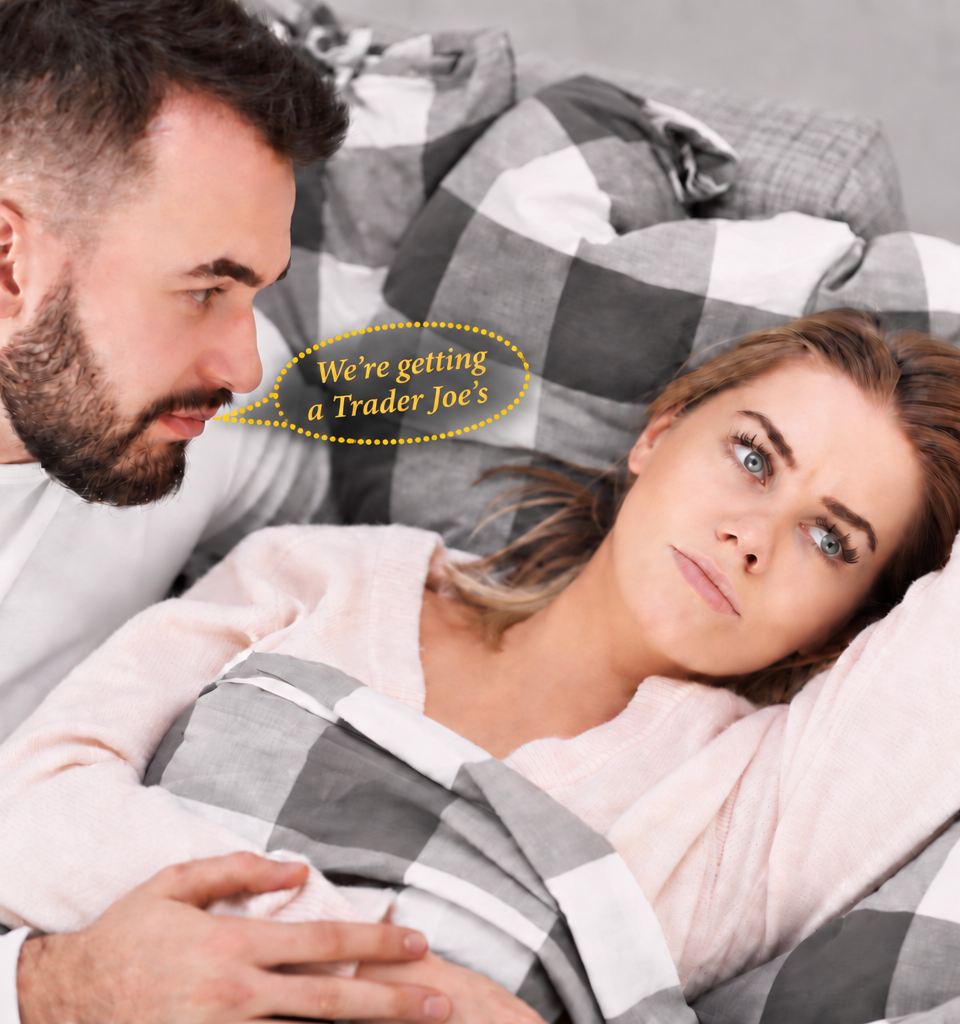 Man leans toward an unimpressed woman in bed, with a dotted gold speech bubble reading “We’re getting a Trader Joe’s.”